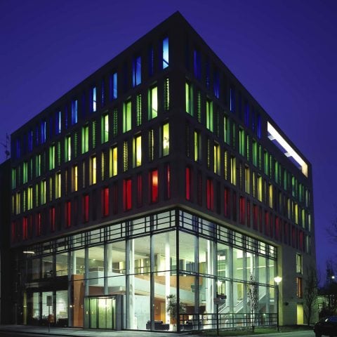 Ein eckiges Gebäude bei Nacht, das hell in den Farben weiß, rot, gelb, grün und blau erleuchtet ist.